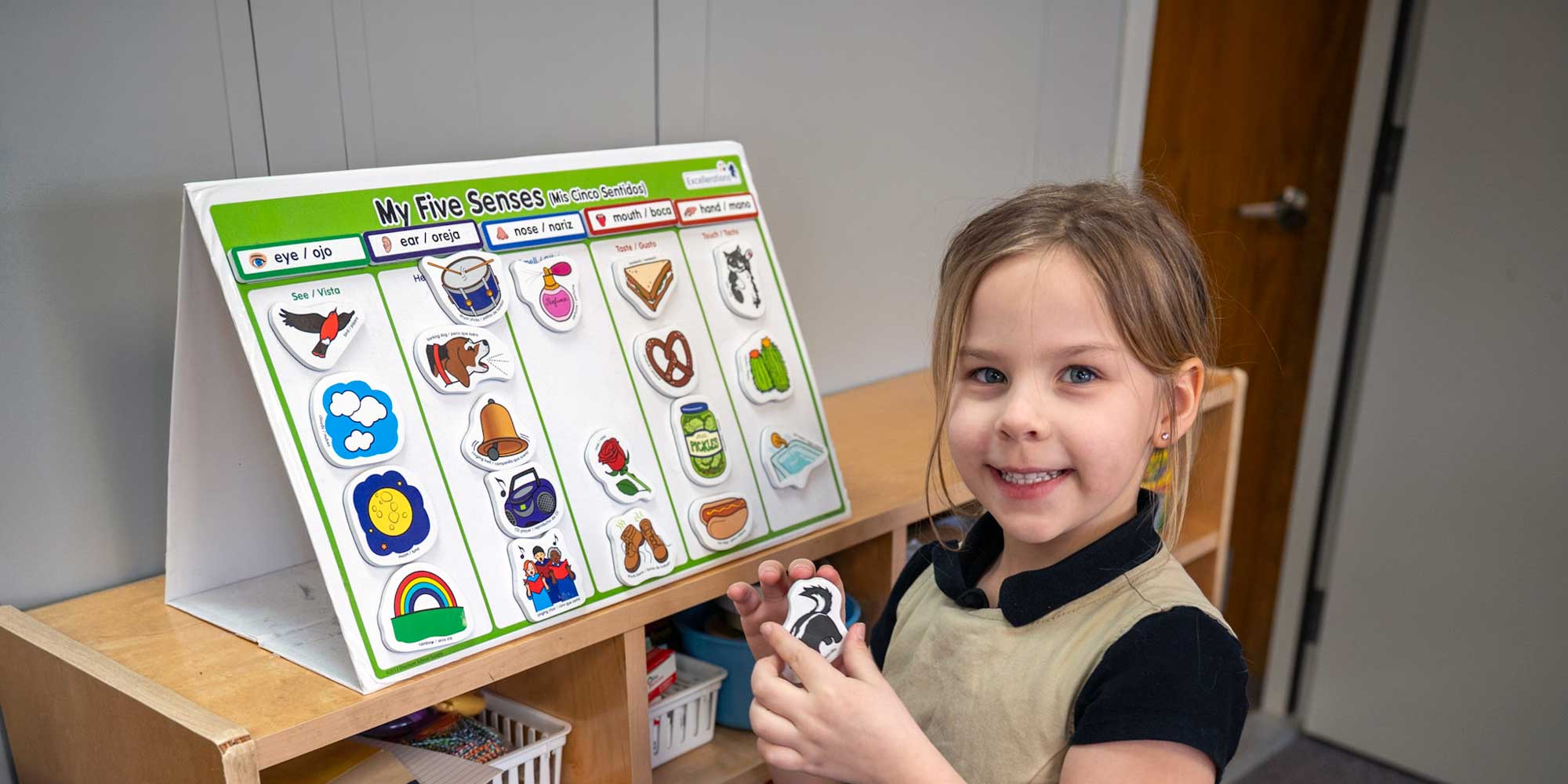 Smiling student holding a picture magnet beside a My Five Senses learning board.