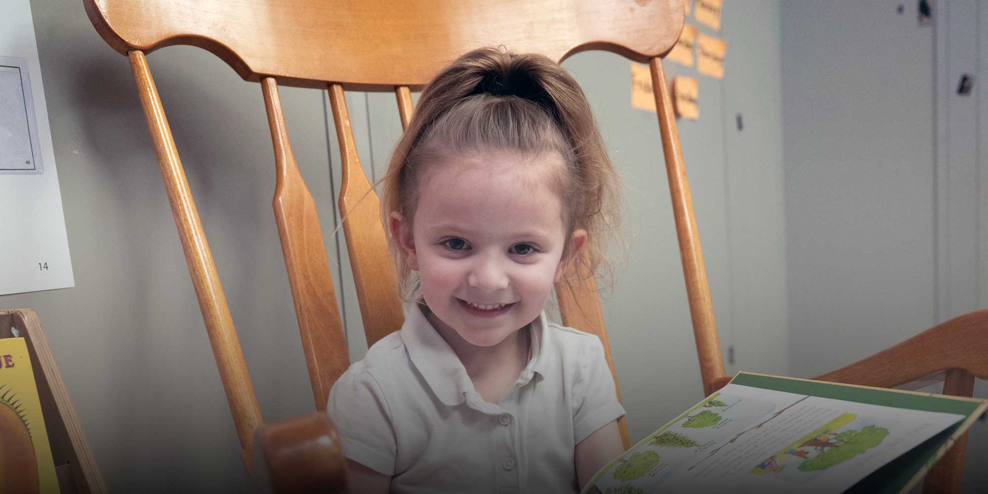 Smiling young student holding an open book in a wooden rocking chair.