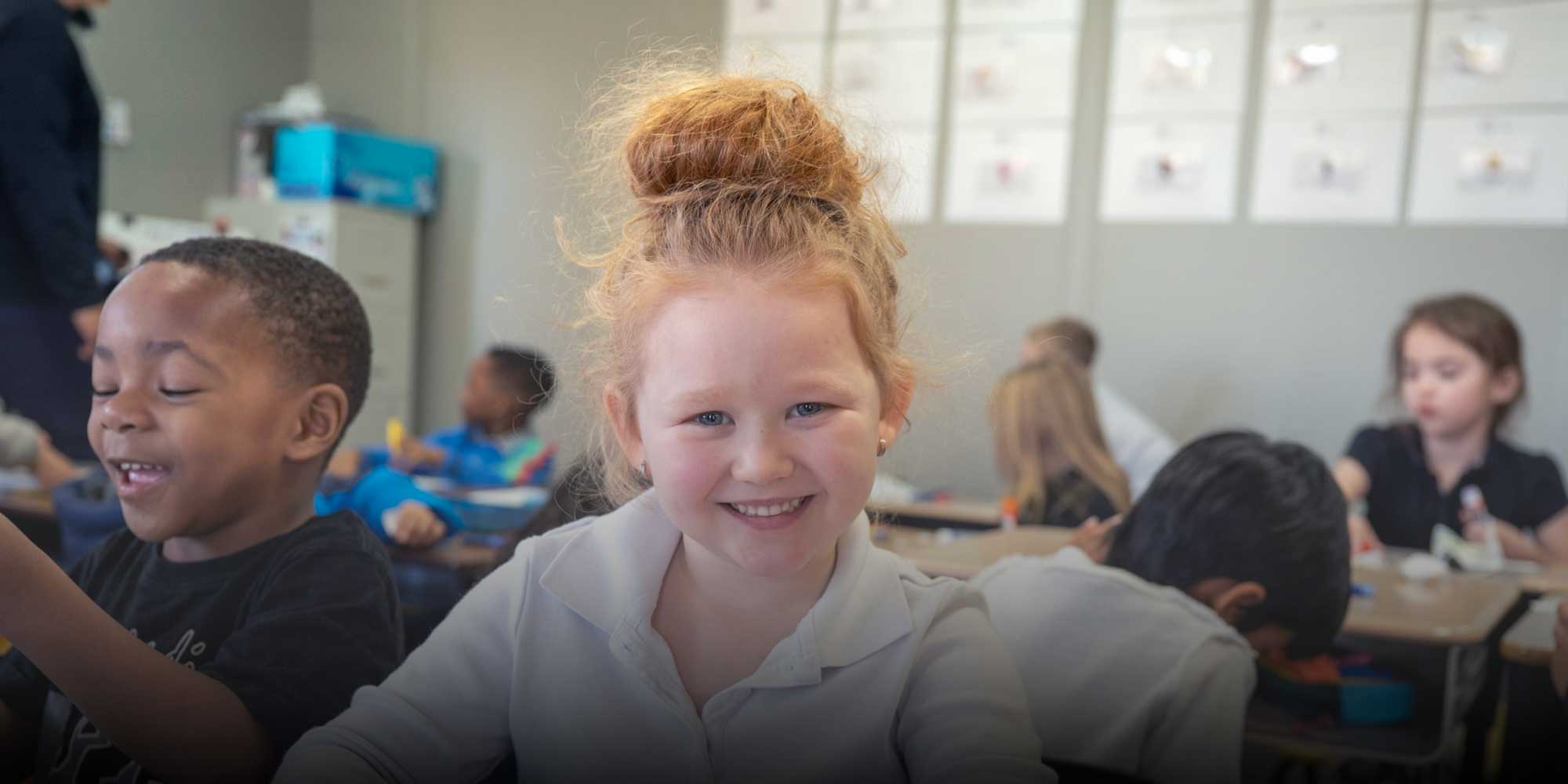 Smiling student at her desk in a busy classroom.