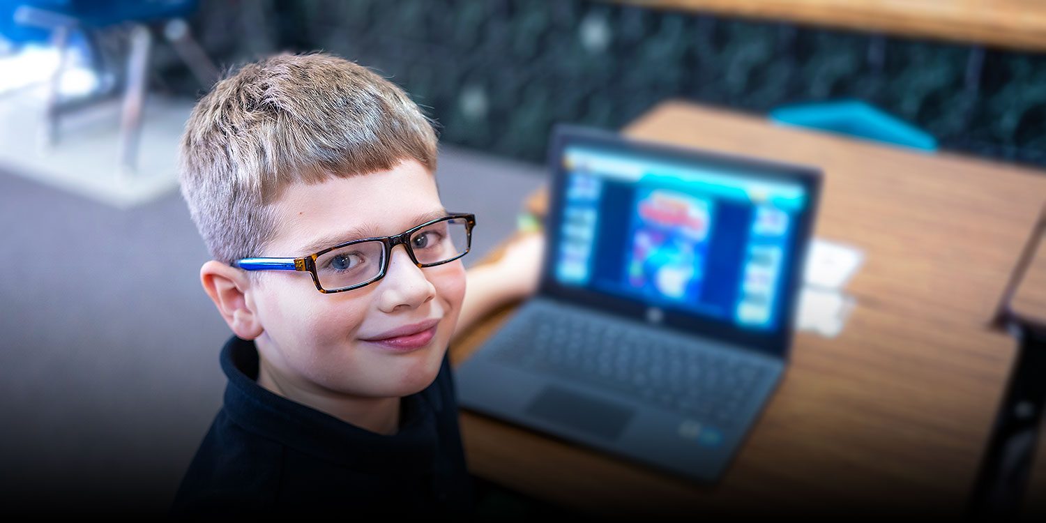 In class, a smiling student sits in front of a laptop.